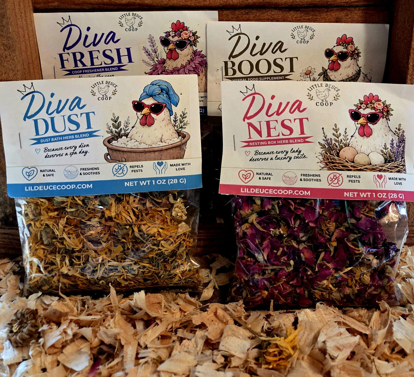 Four bags of Lil' Deuce Coop dried herbs and flowers arranged on wood with shavings. The packages are Diva Fresh, Diva Boost, Diva Dust, and Diva Nest, each showing illustrated hens and different dried botanical contents in clear bags with white labels and colored borders.