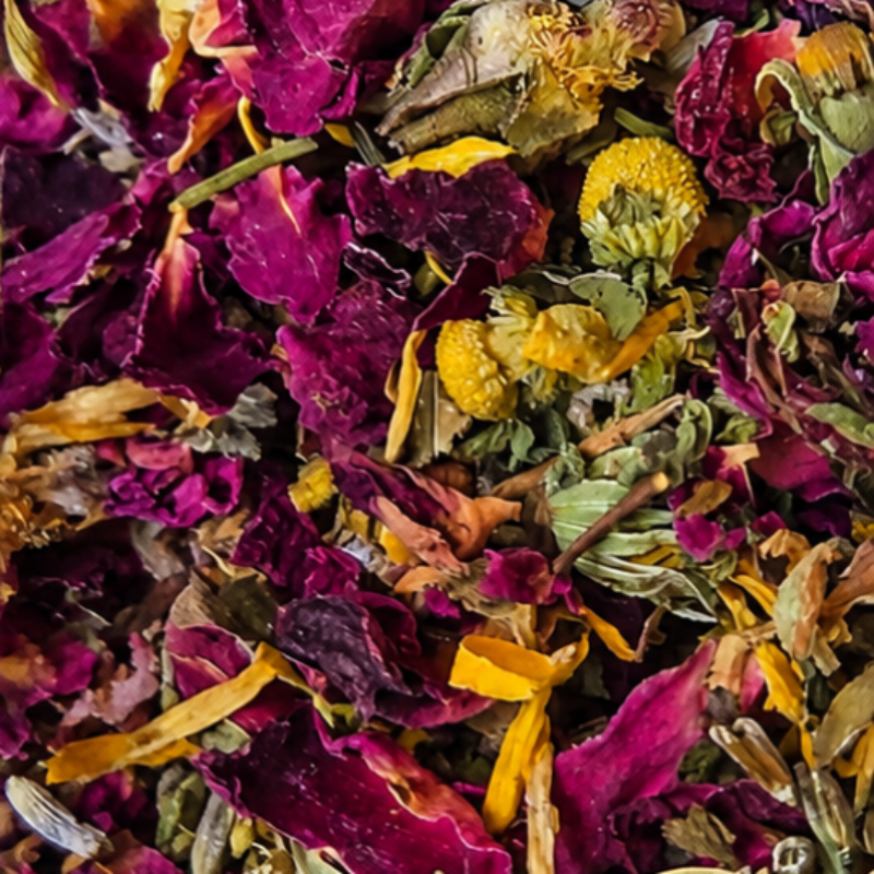 A close-up of dried flower petals and botanicals mixed together, featuring prominent deep purple and magenta rose petals with yellow chamomile flowers and green leaves.
